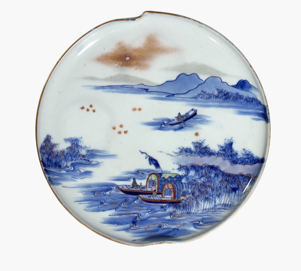 Piatto Imari in porcellana bianca e blu con un paesaggio di acqua, montagne e imbarcazioni. Giappone, fine XIX-XX secolo